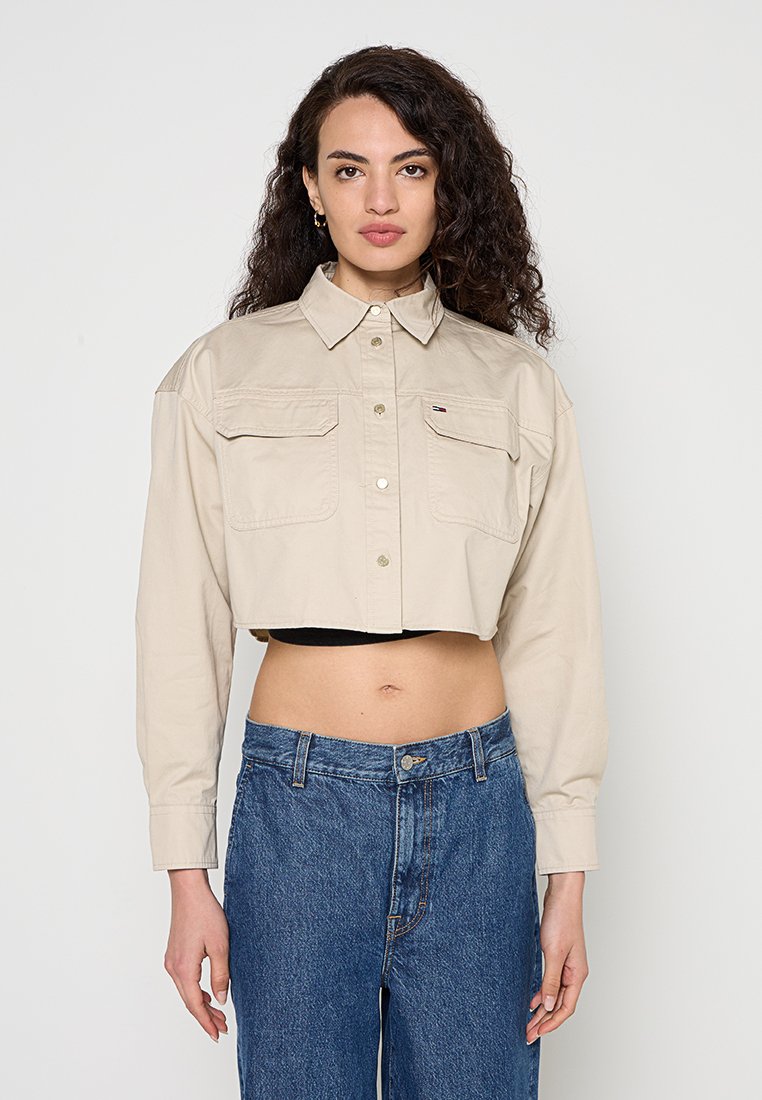 Frau mit lockigem Haar, die eine beige, kurz geschnittene Jacke mit Knöpfen und hoch taillierte blaue Jeans trägt, steht vor einem schlichten Hintergrund.