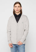 Grauer Baumwoll-Cardigan mit V-Ausschnitt, Knopfverschluss, gerippten Bündchen und kleinem Logo. Getragen über einem schwarzen Hemd mit Kragen und blue Jeans.