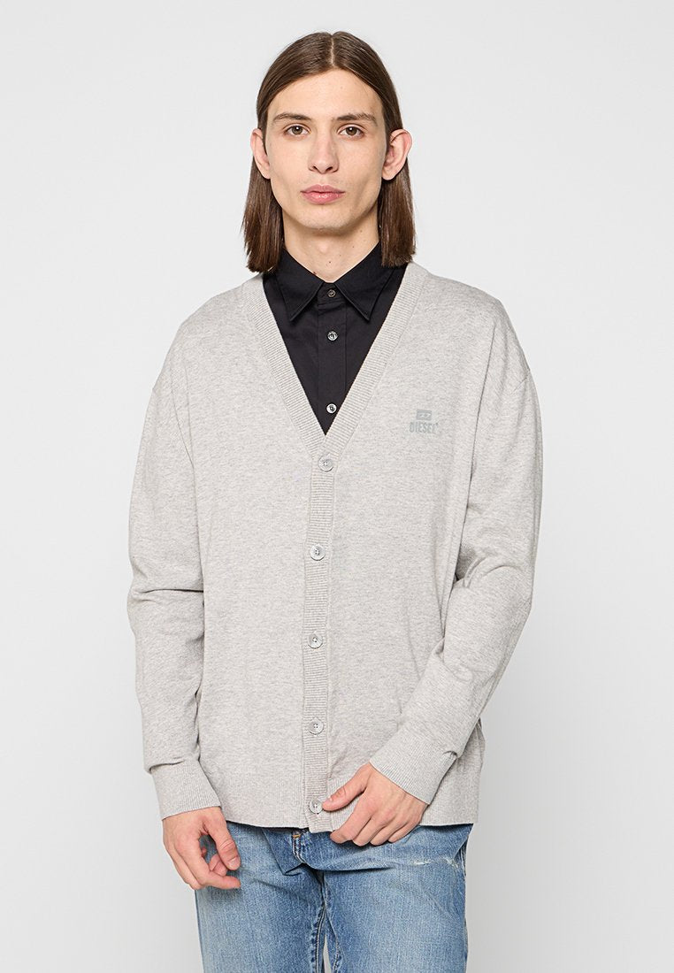 Grauer Baumwoll-Cardigan mit V-Ausschnitt, Knopfverschluss, gerippten Bündchen und kleinem Logo. Getragen über einem schwarzen Hemd mit Kragen und blue Jeans.