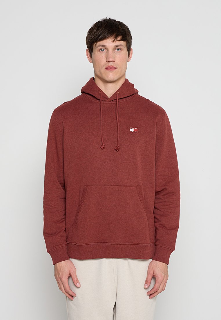 Mann trägt einen rostfarbenen Hoodie mit Fronttasche und kleinem Logo auf der Brust, steht vor neutralem Hintergrund.
