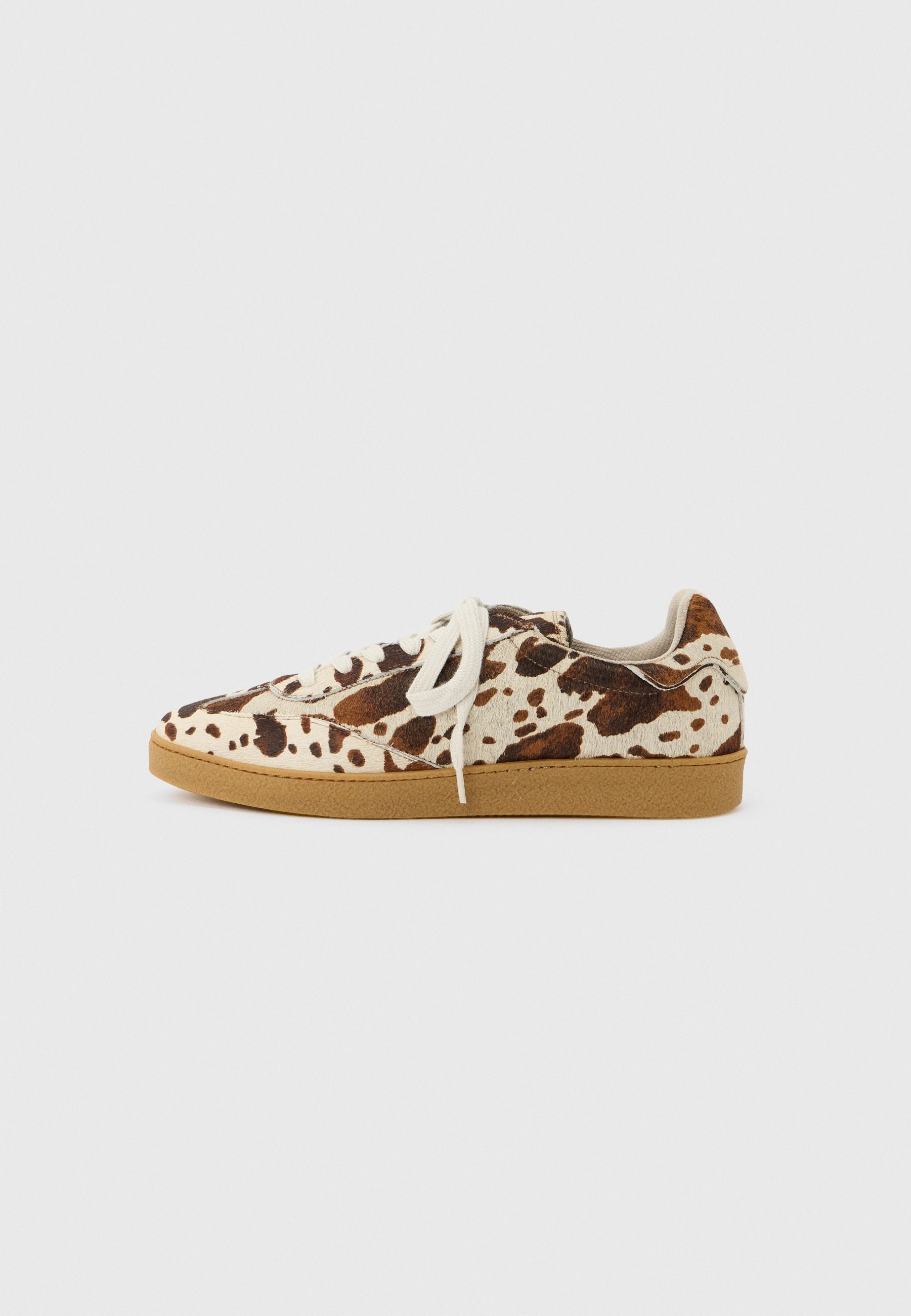 Niedrig geschnittener Sneaker mit weißem und braunem Kuhfellmuster, weißen Schnürsenkeln und einer Sohle aus Gummilaufsohle auf weißem Hintergrund.