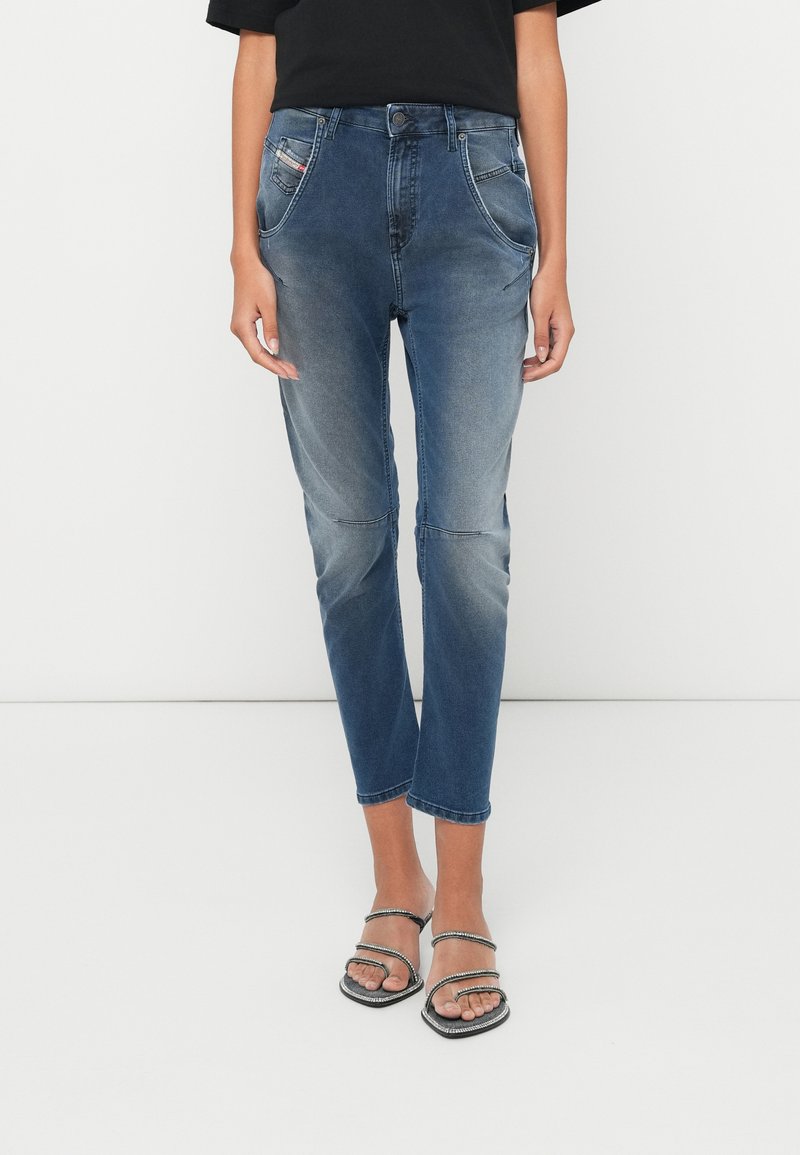 Person in verblassten blauen, hüftlangen Jeans mit geschwungenen Fronttaschen und schwarzen Sandalen mit Riemchen, steht vor einem weißen Hintergrund.