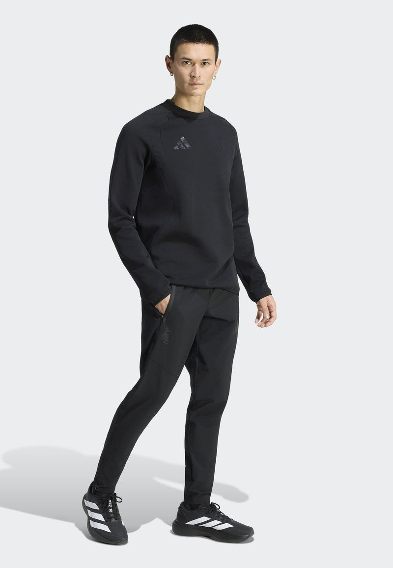Mann trägt schwarzen Adidas-Sweatshirt, schwarze Jogginghose und schwarze Adidas-Sneaker mit weißen Streifen, steht vor einem schlichten Hintergrund.