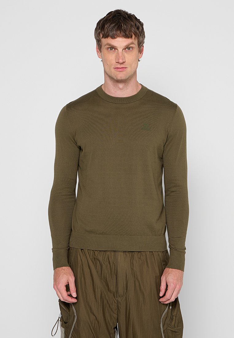 Mann steht da und trägt einen olivgrünen Langarmpullover und passende Hosen mit Taschen, wobei er frontal vor einem einfachen hellen Hintergrund steht.