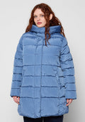 Frau mit langem roten Haar, die eine hellblaue, gesteppte Steppjacke mit hohem Kragen trägt und vor einem schlichten, hellen Hintergrund steht.