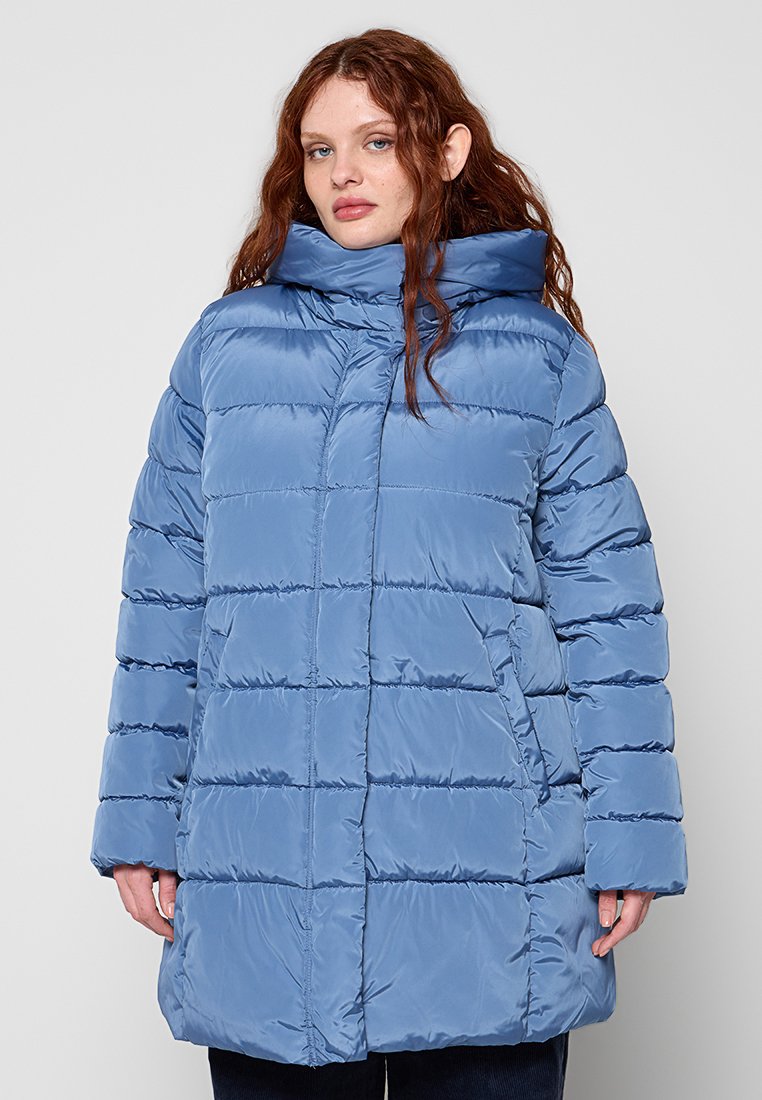 Frau mit langem roten Haar, die eine hellblaue, gesteppte Steppjacke mit hohem Kragen trägt und vor einem schlichten, hellen Hintergrund steht.