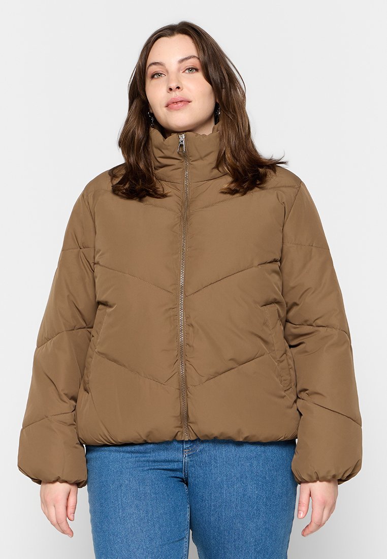 Eine Frau trägt eine braune Steppjacke mit hohem Kragen und blue Jeans und steht vor einem schlichten hellen Hintergrund.