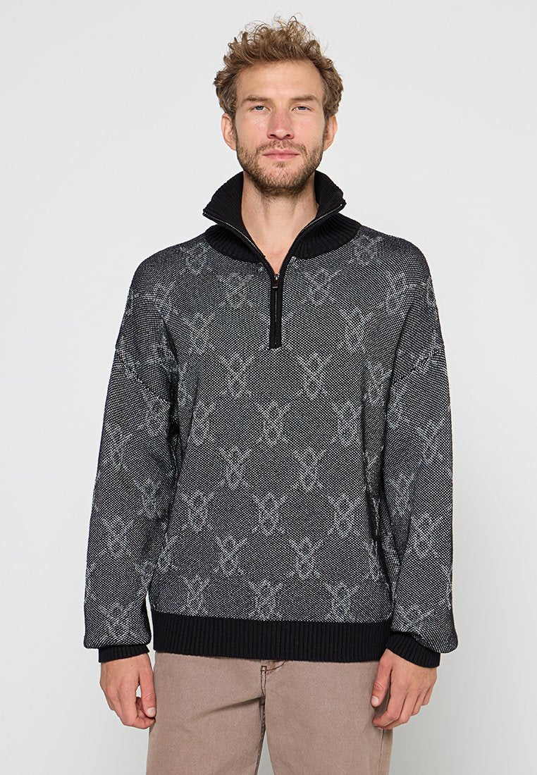 Mann, der einen schwarz-grau gemusterten Halbreißverschluss-Pullover und beige Hosen trägt, steht vor einem schlichten Hintergrund.