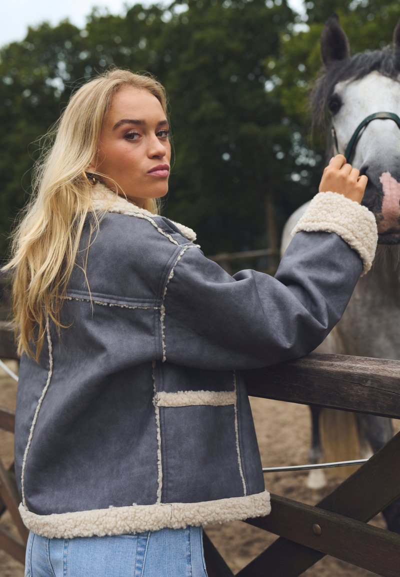 Graue Kunstlederjacke mit weißem Fleece-Futter und -Ärmeln, mit kastigem Design und Taschen vorne, getragen von einer Person in der Nähe eines Pferdes.