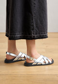 Silberne Sandalen mit Kreuzriemen und flacher schwarzer Sohle sowie einer verstellbaren Schnalle. Sie zeichnen sich durch eine glatte Textur und klare Linien aus.