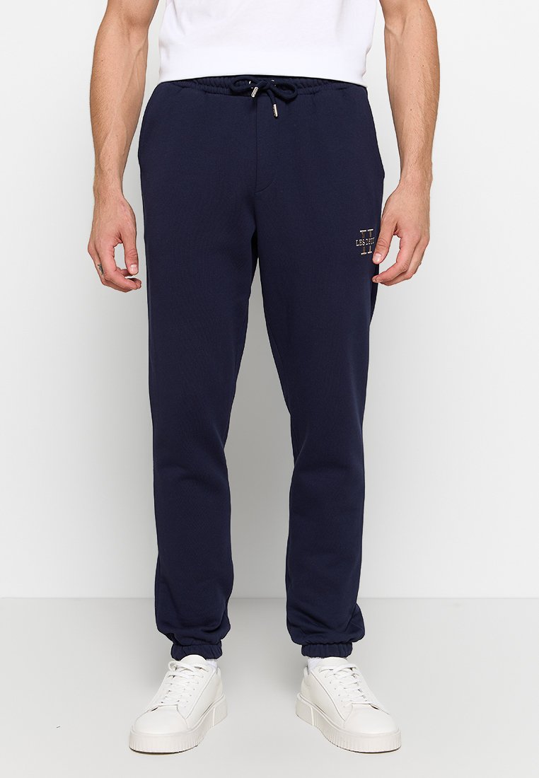Mann trägt marineblaue Jogginghosen mit elastischen Bündchen und weiße Sneaker, stehend vor einem schlichten hellen Hintergrund.