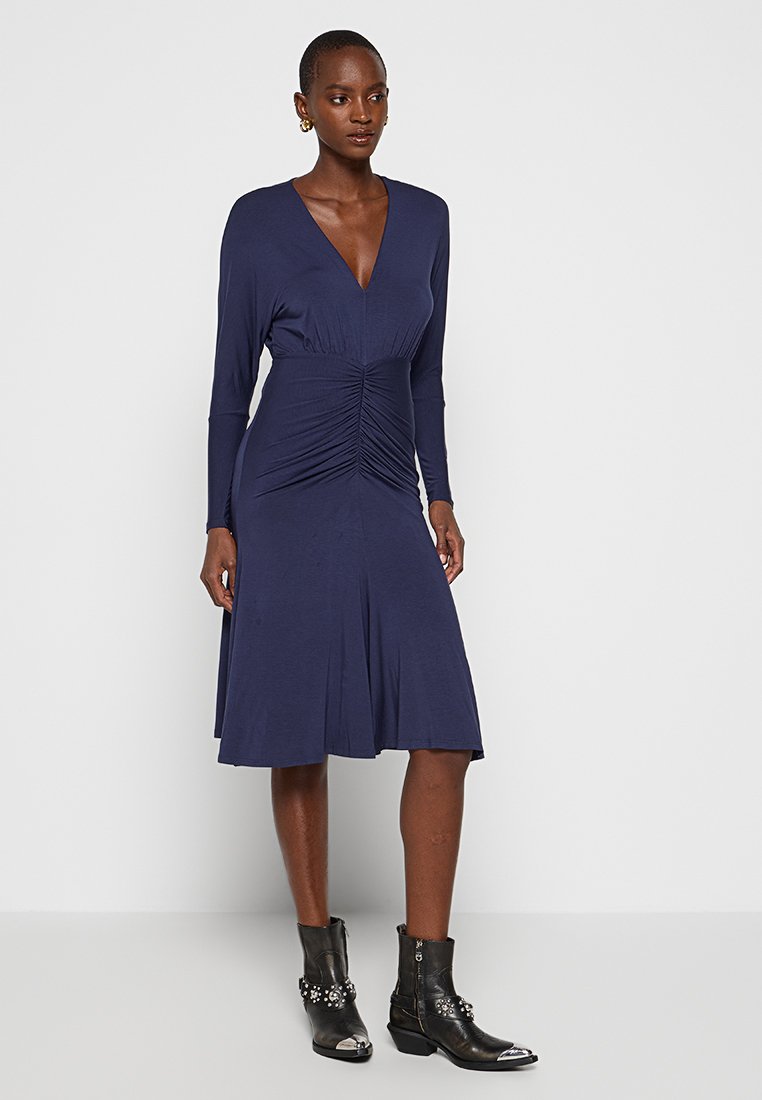 Marineblaue Langarmkleid mit gerafftem Oberteil, V-Ausschnitt, fließendem Rock und kombiniert mit schwarzen, verzierten Knöchelstiefeln. Glatte Stofftextur.