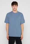 Junger Mann mit kurzen lockigen braunen Haaren, der ein schlichtes blaues T-Shirt mit kurzen Ärmeln und schwarze Hose trägt, steht vor einem hellgrauen Hintergrund.