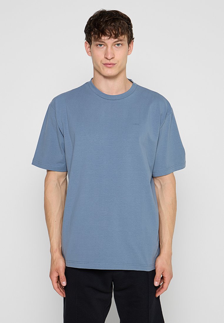 Junger Mann mit kurzen lockigen braunen Haaren, der ein schlichtes blaues T-Shirt mit kurzen Ärmeln und schwarze Hose trägt, steht vor einem hellgrauen Hintergrund.