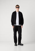 Schwarze Bomberjacke, weißes T-Shirt, schwarze Cargohosen und schwarze Schuhe. Modell trägt Sonnenbrille. Glatte Textur, lässiges Design, keine Muster.