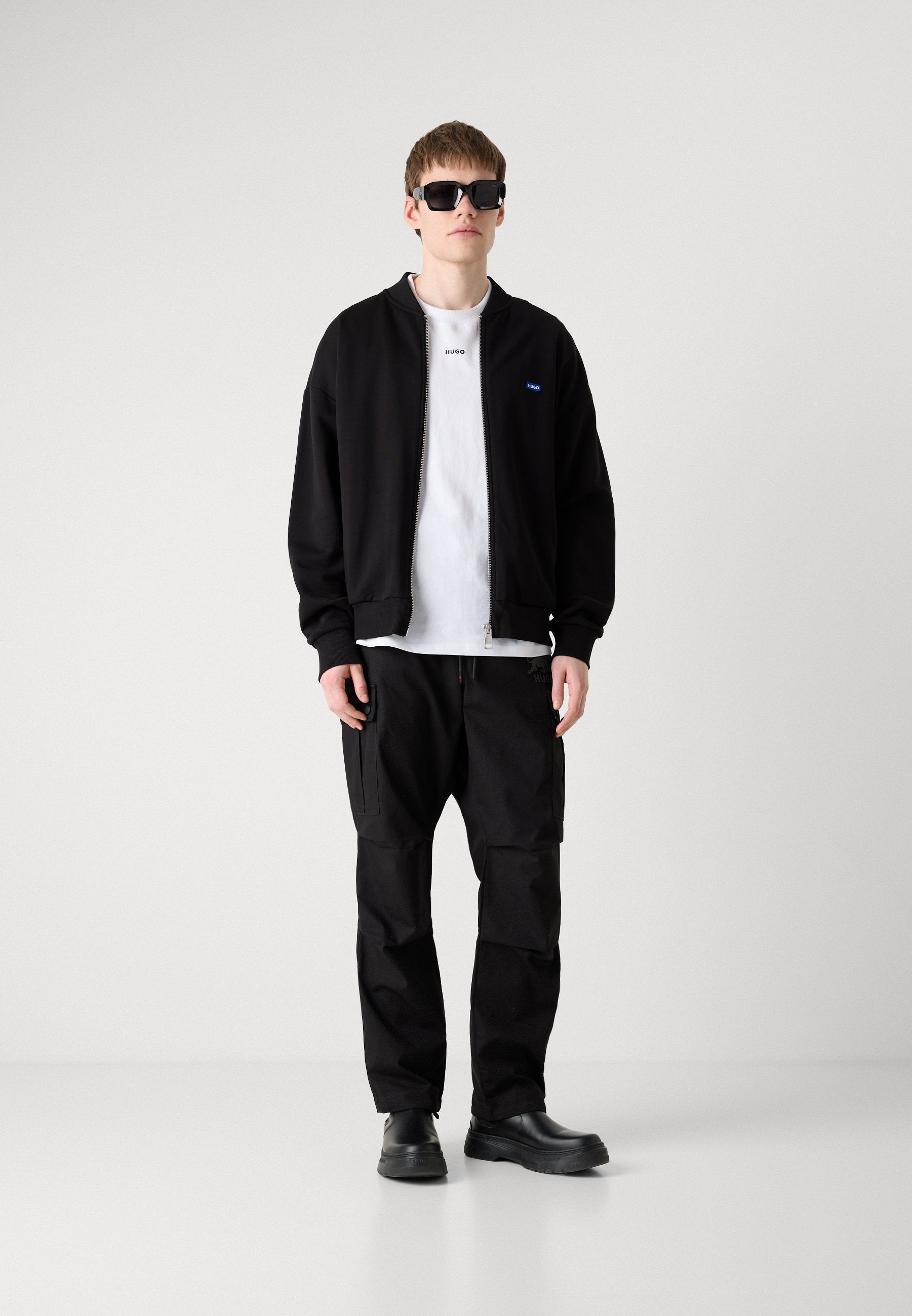 Schwarze Bomberjacke, weißes T-Shirt, schwarze Cargohosen und schwarze Schuhe. Modell trägt Sonnenbrille. Glatte Textur, lässiges Design, keine Muster.