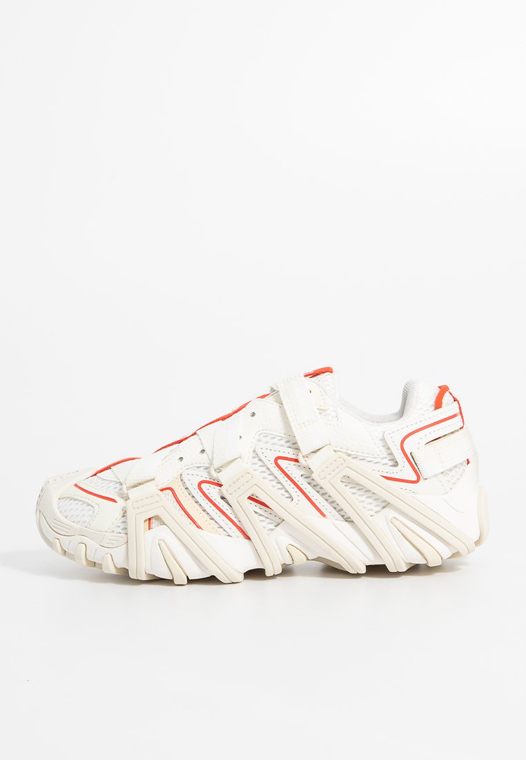 Weißes und beige Sport Schuh mit roten Akzenten, Netzpaneelen und mehreren Riemen über der Oberseite und den Seiten auf weißem Hintergrund.