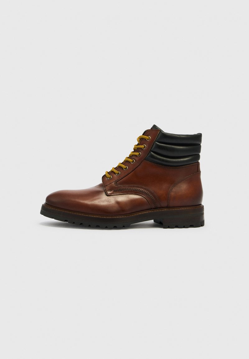 Brauner Leder-Knöchelstiefel mit schwarzem gepolstertem Kragen, gelben und braunen Schnürsenkeln sowie robuster schwarzer Sohle auf einfarbigem Hintergrund.