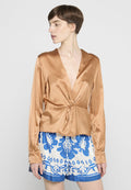 Bronzefarbene Satin-Wickelbluse mit langen Ärmeln, tiefem V-Ausschnitt und gebundenem Vorderteil, kombiniert mit blauen und weißen gemusterten Shorts mit floralen Motiven.