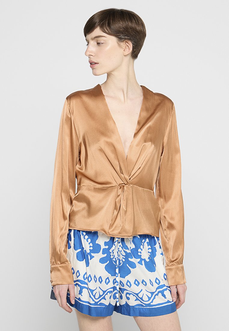 Bronzefarbene Satin-Wickelbluse mit langen Ärmeln, tiefem V-Ausschnitt und gebundenem Vorderteil, kombiniert mit blauen und weißen gemusterten Shorts mit floralen Motiven.