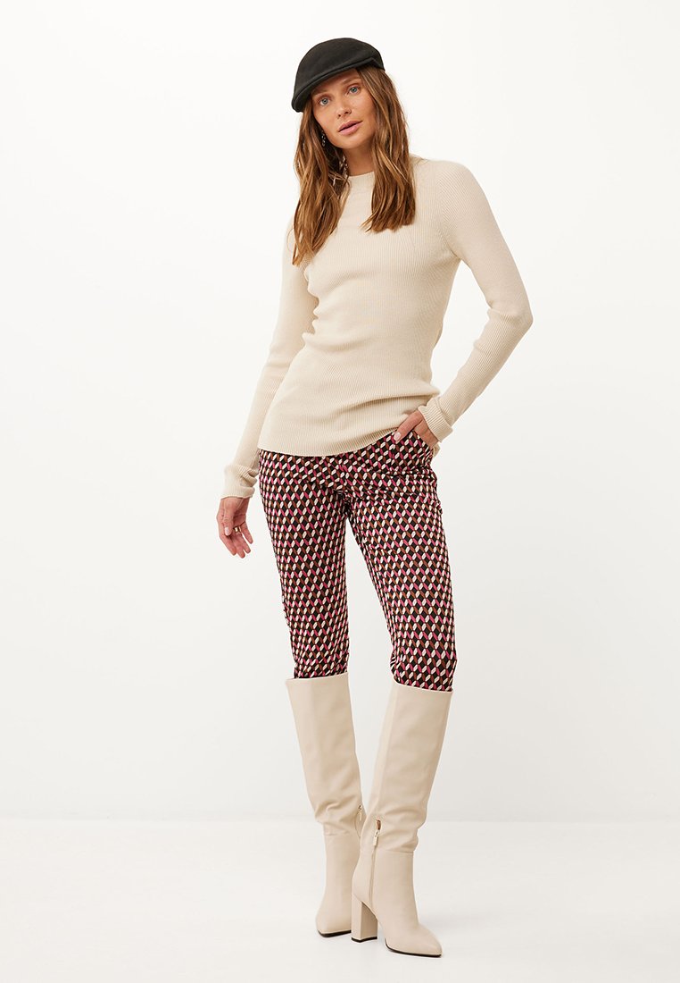 Beige geripptes Langarmoberteil, gemusterte rot-weiße eng anliegende Hose und knielange beige Stiefel mit Absatz. Der Hut ist schwarz und setzt einen Kontrast.