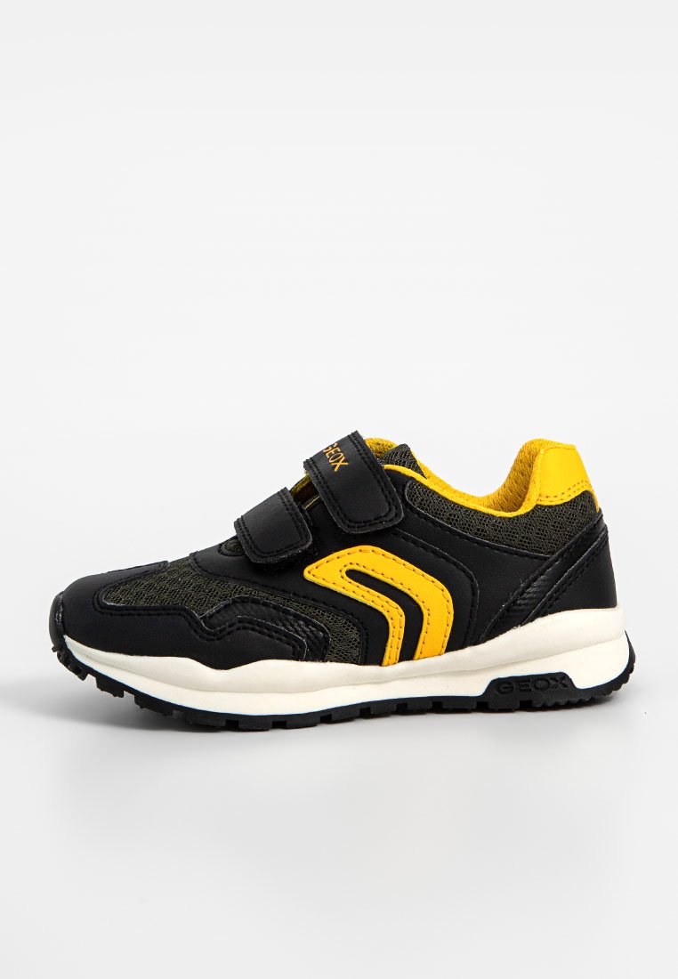 Schwarze Sneakers mit gelben Akzenten, ausgestattet mit einem Klettverschluss, Netzpaneelen, gepolstertem Kragen und einer strukturierten Gummisohle für besseren Halt.