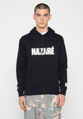 Junger Mann mit kurzen Haaren, der einen schwarzen "NAZARÉ" Hoodie und hellblaue, beige und graue Camouflage-Hosen trägt, steht vor einem weißen Hintergrund.