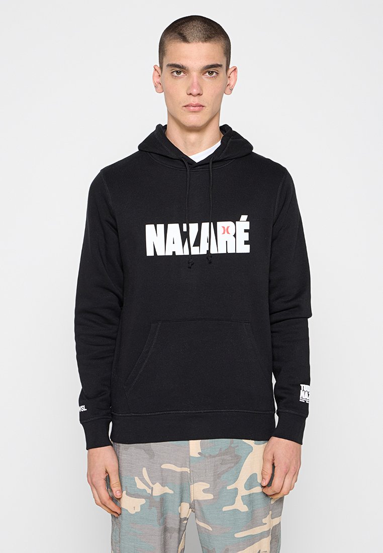 Junger Mann mit kurzen Haaren, der einen schwarzen "NAZARÉ" Hoodie und hellblaue, beige und graue Camouflage-Hosen trägt, steht vor einem weißen Hintergrund.