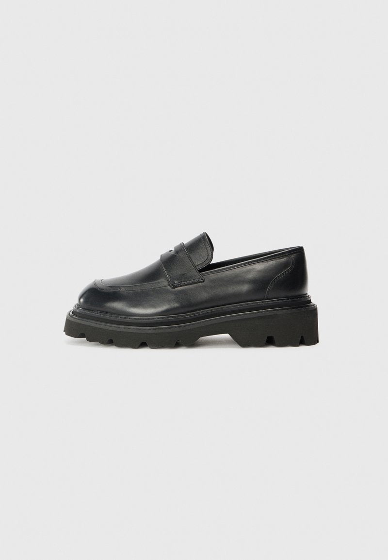 Schwarze Lederloafer mit runder Zehenform, ausgestattet mit einer niedrigen, klobigen Gummisohle mit einem gemusterten Profil und einem gefalteten Riemen auf dem Schaft.
