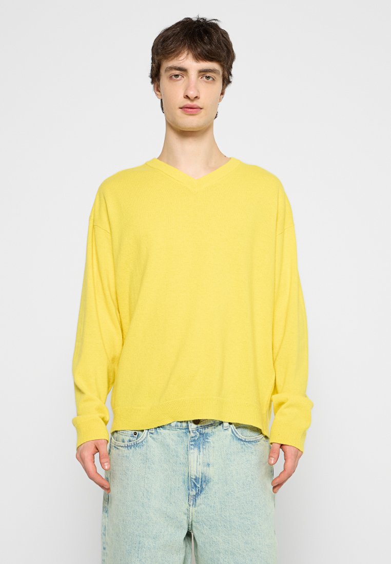 Gelber V-Ausschnitt-Pullover mit langen Ärmeln, lockere Passform, aus weichem Material; kombiniert mit hellblauen, verwaschenen Denim-Jeans.