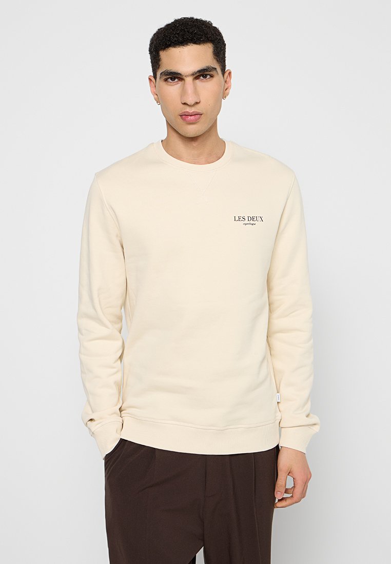 Helle beige Sweatshirt mit langen Ärmeln, Rundhalsausschnitt und gerippten Bündchen. Mit minimalistischem schwarzem Text "LES DEUX" auf der Brust. Weicher Stoff.