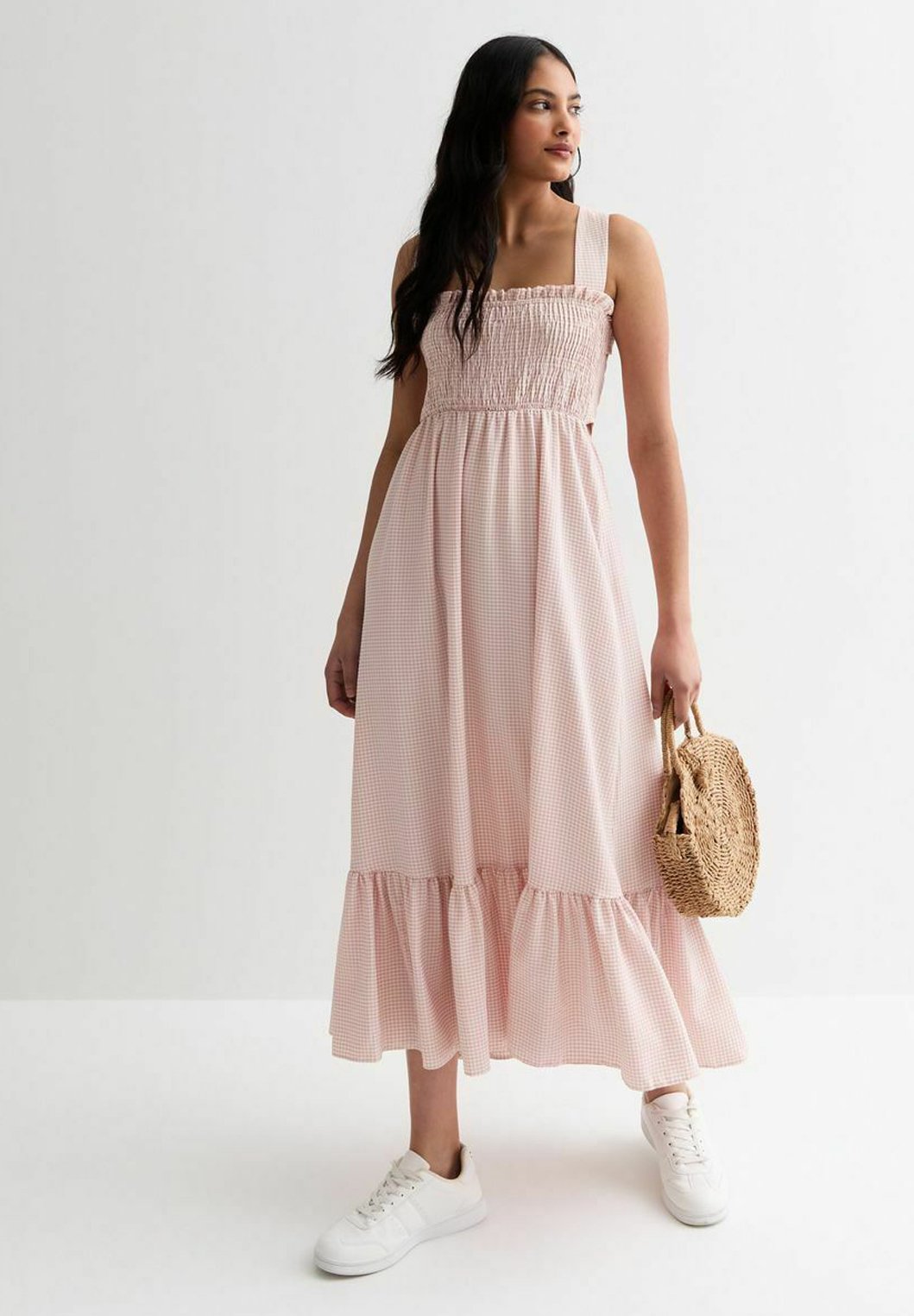 Rosa Karomuster-Maxikleid mit elastischem Oberteil, breiten Trägern und Rüschensaum. Kombiniert mit weißen Sneakers und einer kleinen geflochtenen Handtasche.