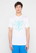 Junger Mann trägt ein weißes Nike-T-Shirt mit blauem Flamingo und den Buchstaben "A R" als Design sowie blaue Sportshorts, steht vor neutralem Hintergrund.