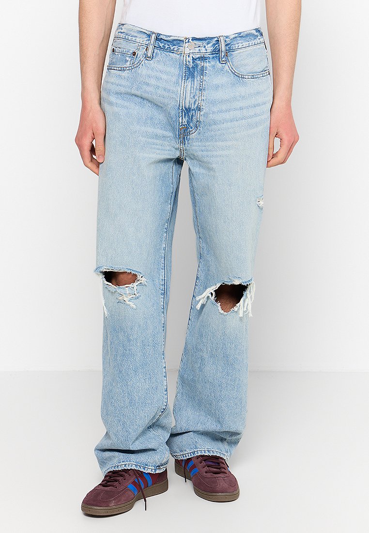 Helle blaue Ripped-Jeans mit fransigen Kanten, geradem Schnitt, fünf Taschen und Metallbeschlägen. Getragen mit dunklen Schuhen mit Streifen.