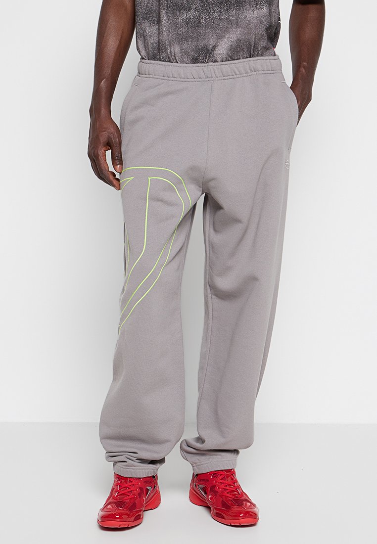 Mann in grauen Jogginghosen mit großem neongelben Design am linken Bein und glänzenden roten Sneakers, der vor weißem Hintergrund steht.