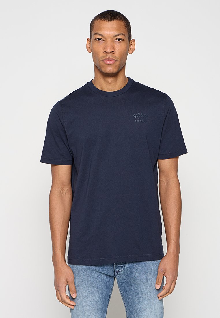 Navyblaues Baumwoll-T-Shirt mit Rundhalsausschnitt und kurzen Ärmeln, versehen mit einem dezenten, aufgedruckten Logo auf der Brust. Kombiniert mit hellblauen Jeans.