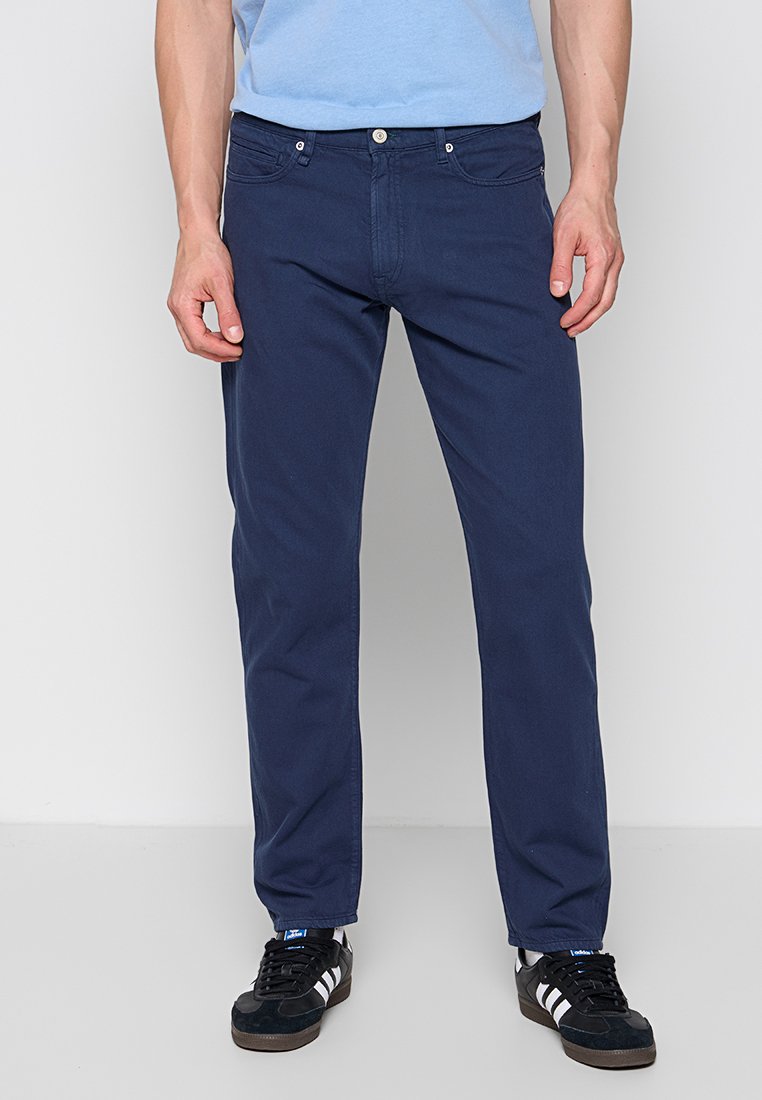 Marineblaue Baumwollhose mit geradem Bein, zwei Fronttaschen und markanten Steppnähten. Getragen mit schwarzen Sneakers.