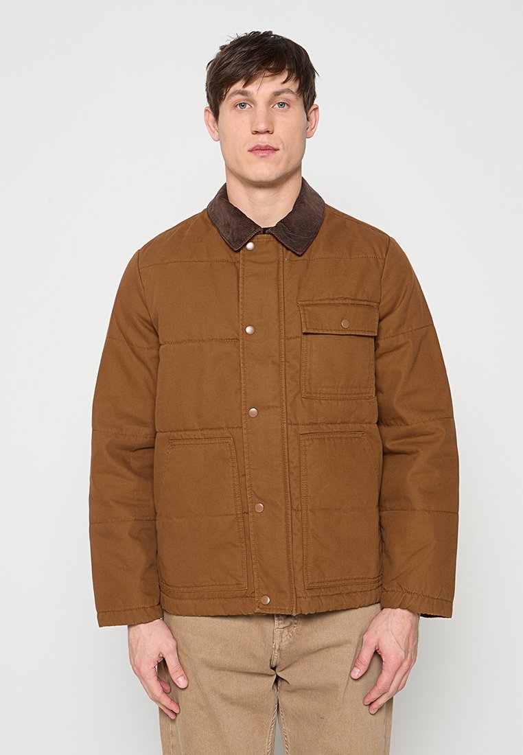 Junger Mann trägt eine braune Steppjacke mit dunklem Kragen und beige Hose, steht vor einem einfarbigen hellen Hintergrund.