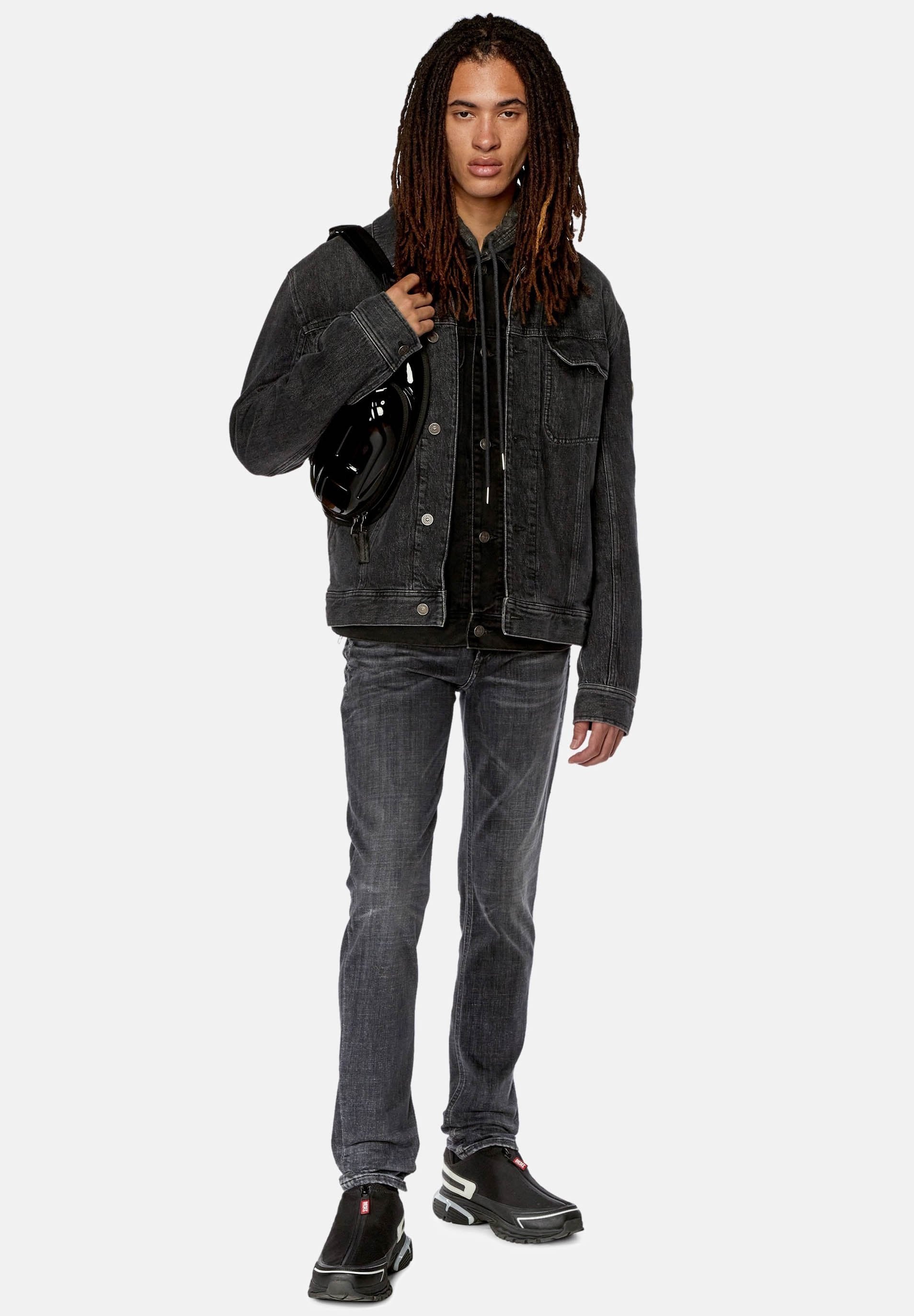 Schwarze Jeansjacke über einem schwarzen Hoodie, graue Jeans und schwarze Schuhe mit weißen Akzenten. Accessories umfassen eine schwarze Tasche. Gerade Passform.