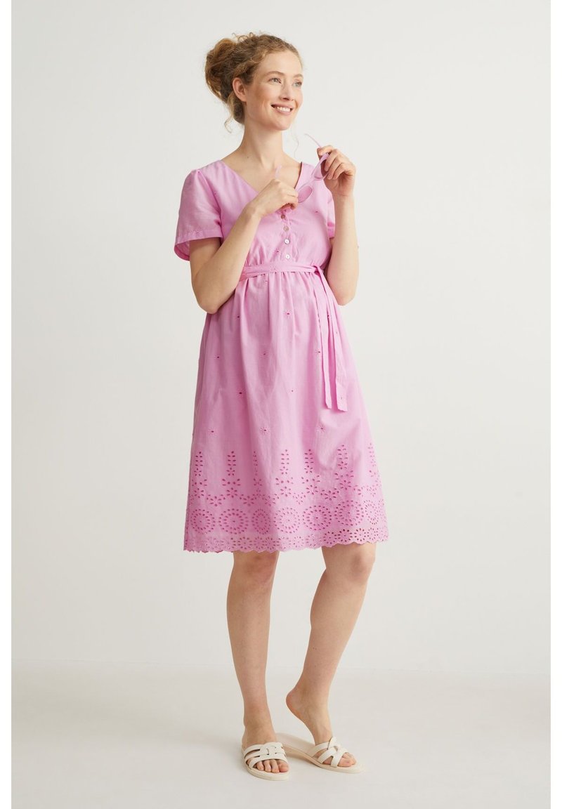 Rosa Baumwollkleid mit kurzen Ärmeln, V-Ausschnitt, Knopfdetails, tailliertem Gürtel und gesticktem Cut-Out-Saum. Kombiniert mit weißen Sandalen.