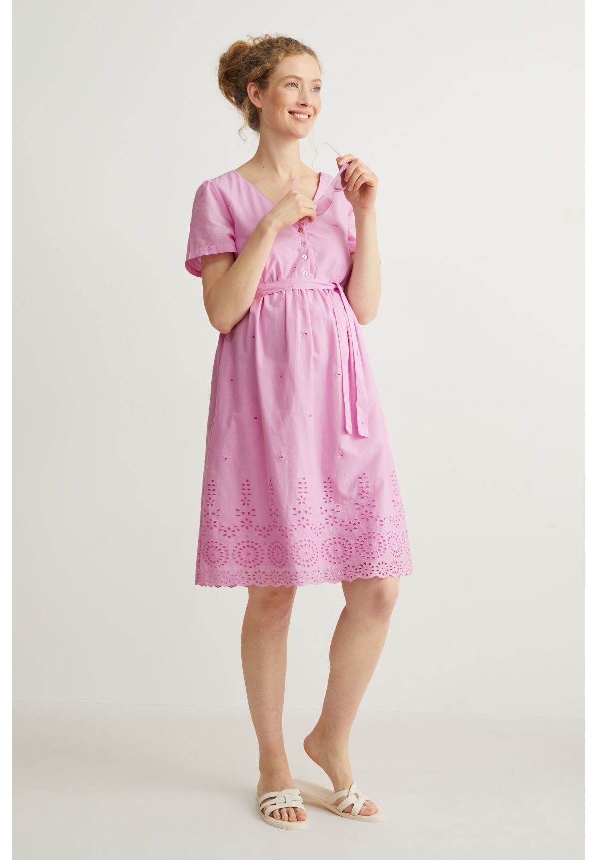 Rosa Baumwollkleid mit kurzen Ärmeln, V-Ausschnitt, Knopfdetails, tailliertem Gürtel und gesticktem Cut-Out-Saum. Kombiniert mit weißen Sandalen.