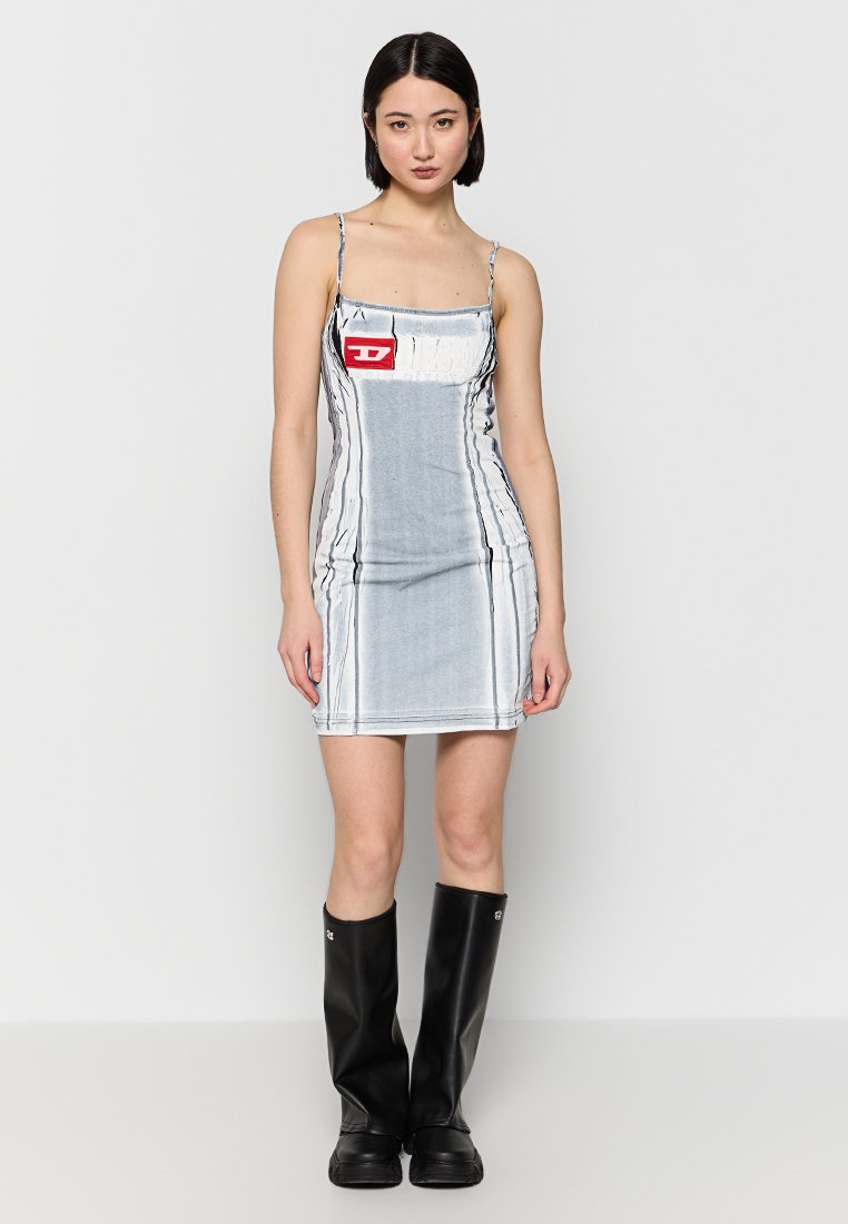 Frau steht in einem enganliegenden grauen und weißen ärmellosen Kleid mit einem roten Logo-Patch und schwarzen kniehohen Stiefeln.