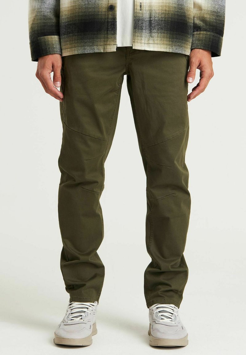 Olivgrüne Cargo-Hosen mit lockerer Passform, ausgestattet mit aufgesetzten Taschen und genähten Akzenten. Getragen mit hellen Sneakers.