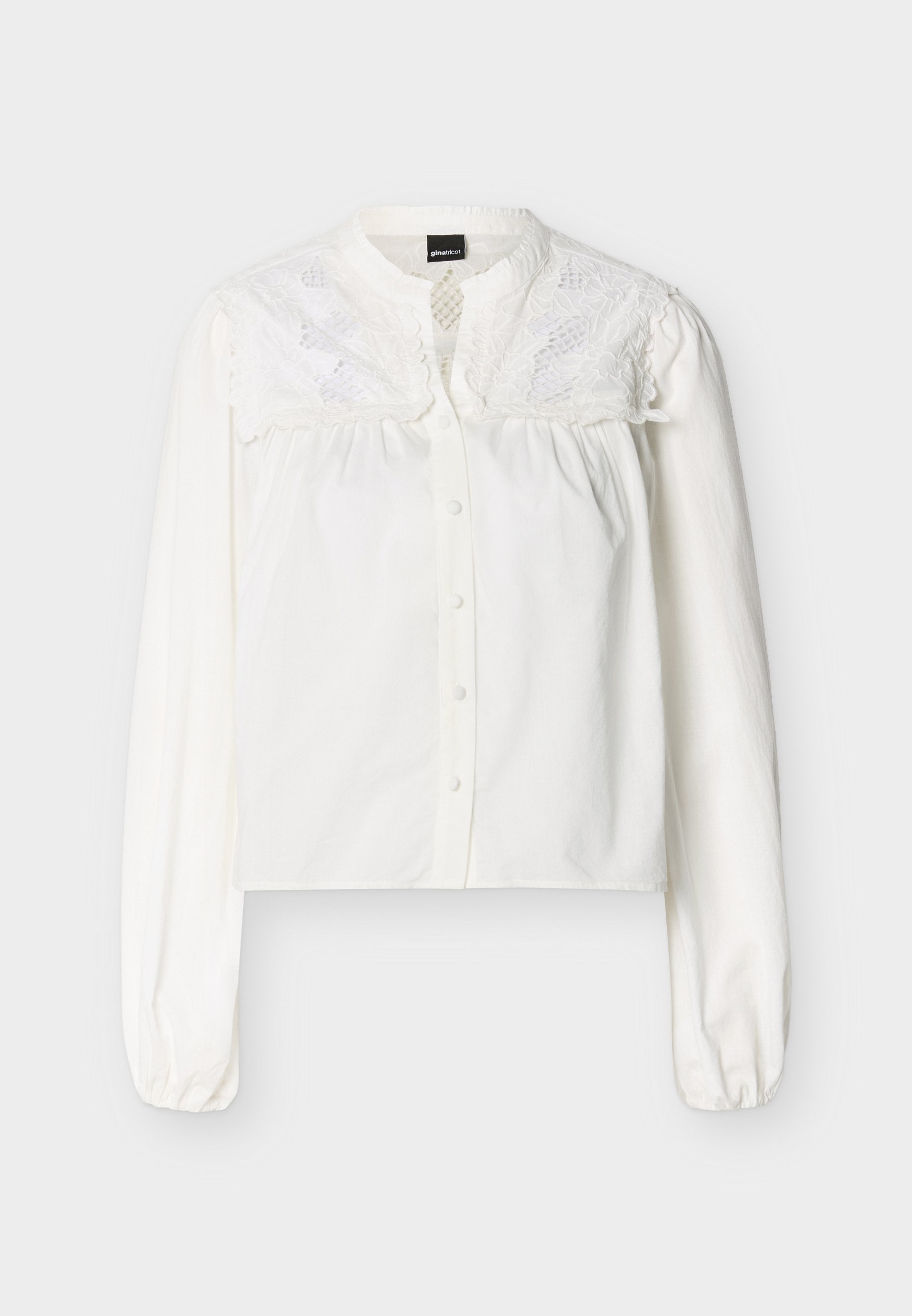 Weiße Bluse mit langen Ärmeln, Frontknöpfen und besticktem floralen Spitzenbesatz im oberen Brust- und Schulterbereich.