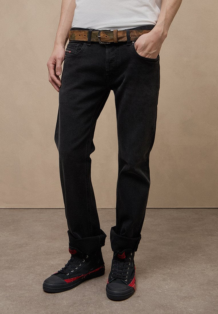 Person trägt schwarze hochgekrempelte Jeans, camouflierter Gürtel, weißes Hemd eingesteckt und schwarze High-Top-Sneaker mit roten Akzenten und steht auf einem grauen Boden.