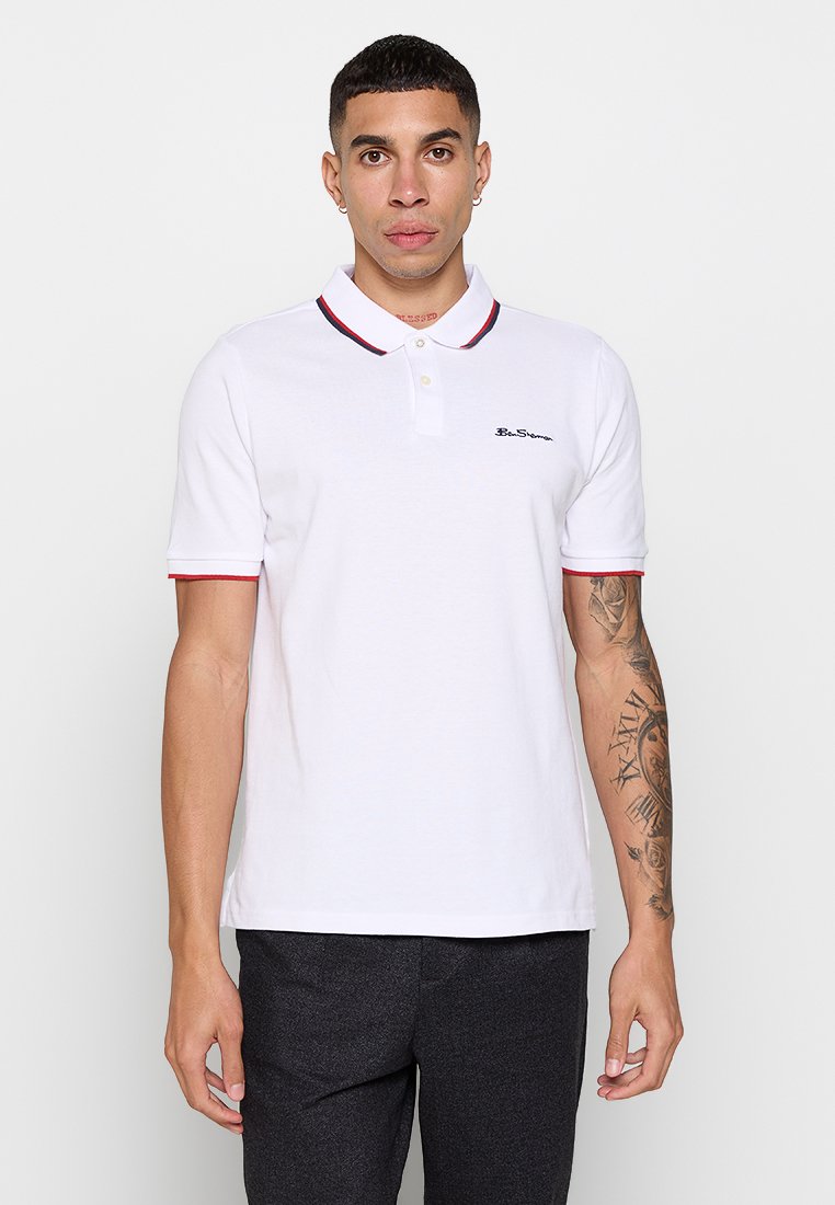 Weißes Polo-Shirt mit kontrastierendem roten und marineblauen Kragenbesatz, kurzen Ärmeln und aufgesticktem Logo auf der linken Brust. Hergestellt aus Baumwolle.