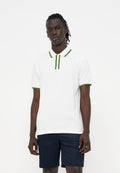 Junger Mann mit dunkler Haut und Dreadlocks, der ein weißes Poloshirt mit grünem Rand und marineblaue Shorts trägt und vor einem einfarbigen Hintergrund steht.