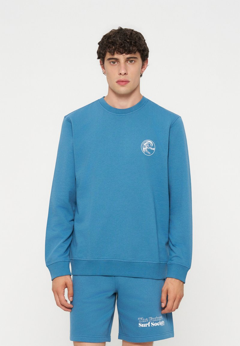 Junger Mann mit lockigem Haar, der einen blauen langärmeligen Sweatshirt und passende Shorts mit weißen Surf-Logos auf Brust und Bein trägt.