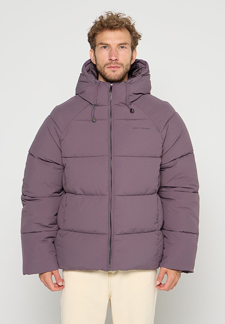 Mann trägt eine lila gepolsterte Kapuzenjacke, die zugezippt ist, und steht entspannt mit den Händen an den Seiten vor einem schlichten hellen Hintergrund.
