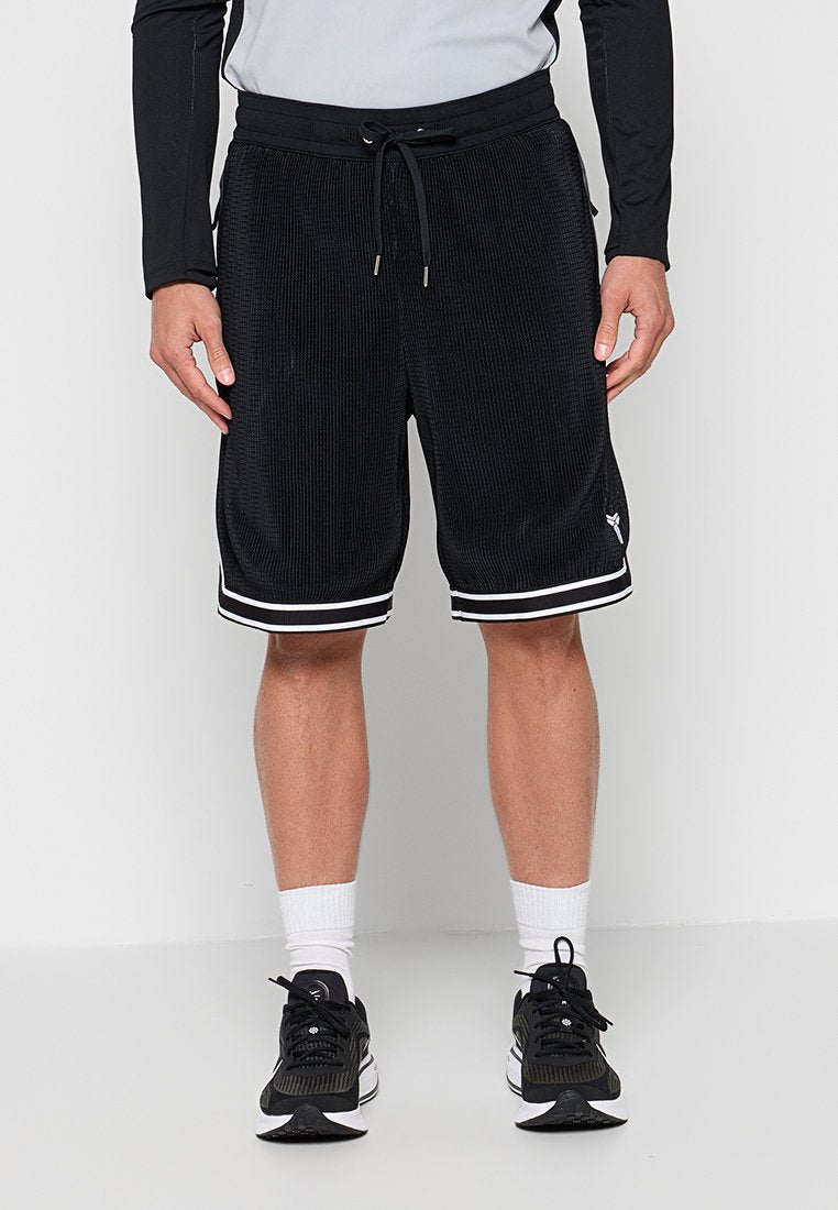 Mann trägt schwarze Basketball-Shorts mit Kordelzug und weißen Streifendetails, weiße Knöchelsocken und schwarze Sportschuhe, steht vor einer weißen Wand.